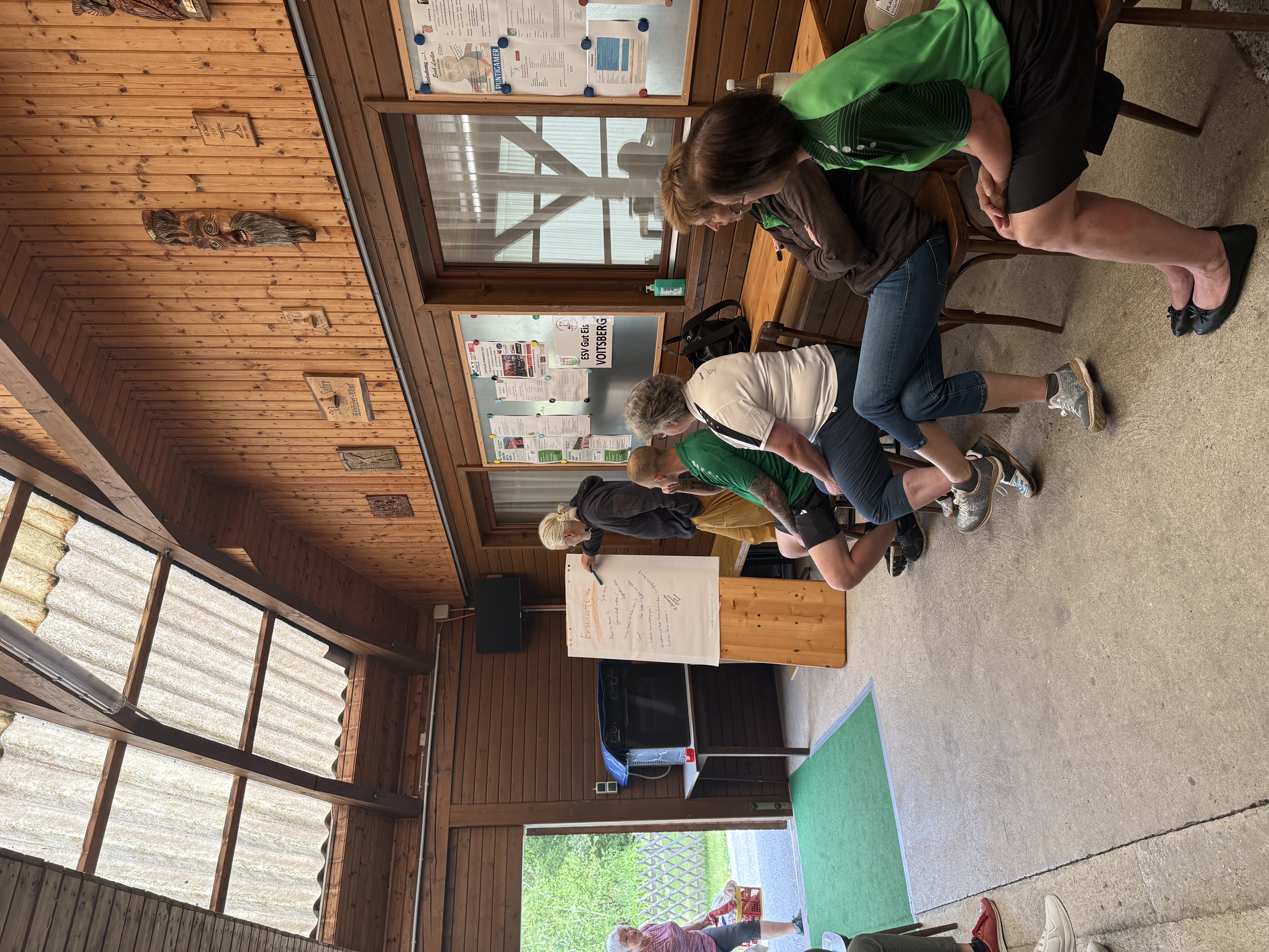 Theorie trifft Praxis: Flipchart-Session mit Trainer Albert Iglseder in der Vereinshütte. Innenansicht einer Holzhütte: Trainer schreibt an ein Flipchart, Teilnehmer*innen sitzen aufmerksam auf Stühlen und Bänken.